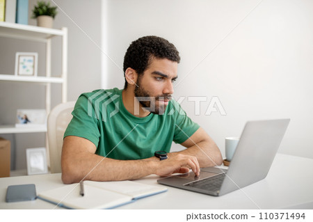 Focused man with beard in green t-shirt working attentively on a laptop in a bright Focused man with beard in green t-shirt working attentively on a laptop in a bright 110371494