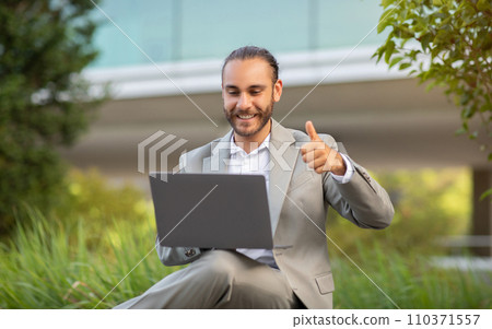 Optimistic young businessman in suit working on laptop outdoors, showing thumb up 110371557