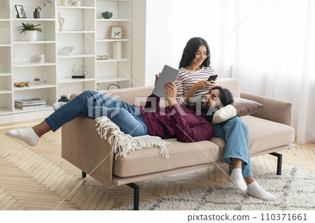 Cheerful eastern man and woman with gadgets chilling at home Cheerful eastern man and woman with gadgets chilling at home 110371661