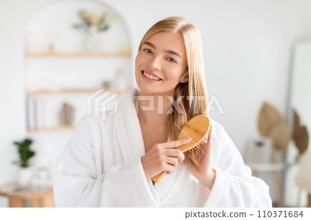 Blonde woman smiling at camera brushing her long hair indoor 110371684