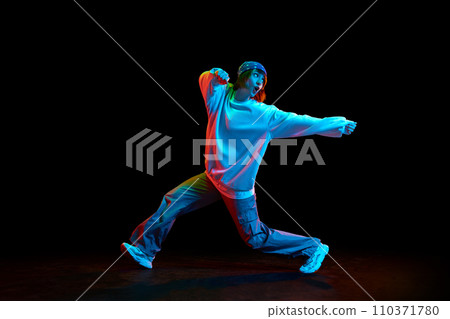 Hip hop culture. Young woman in sportive clothe dancing modern dance styles isolated over black background in neon light Hip hop culture. Young woman in sportive clothe dancing modern dance styles isolated over black background in neon light 110371780