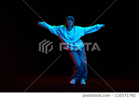 Hip hop culture. Young woman in sportive clothe dancing modern dance styles isolated over black background in neon light Hip hop culture. Young woman in sportive clothe dancing modern dance styles isolated over black background in neon light 110371792