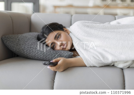 Bored indian woman with remote controller lying on sofa at home Bored indian woman with remote controller lying on sofa at home 110371996