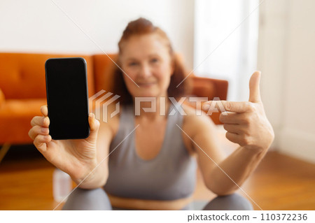 Smiling elderly lady in sportswear pointing at blank smartphone screen 110372236