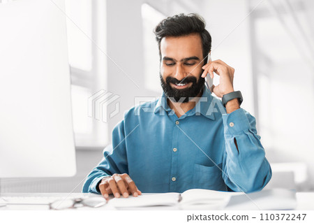 Joyful indian man conversing on phone with planner and glasses Joyful indian man conversing on phone with planner and glasses 110372247