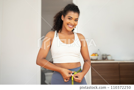 Athletic young woman measuring waist smiling to camera at home Athletic young woman measuring waist smiling to camera at home 110372250