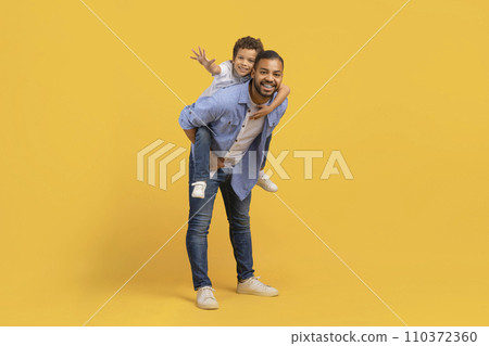 Loving black dad giving his son a piggyback ride over yellow background Loving black dad giving his son a piggyback ride over yellow background 110372360