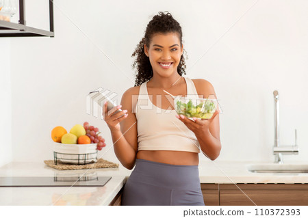 Happy sporty woman holds smartphone and fresh vegetable salad indoor 110372393