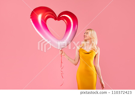 Joyful blonde woman with two-tone heart balloon on pink background Joyful blonde woman with two-tone heart balloon on pink background 110372423