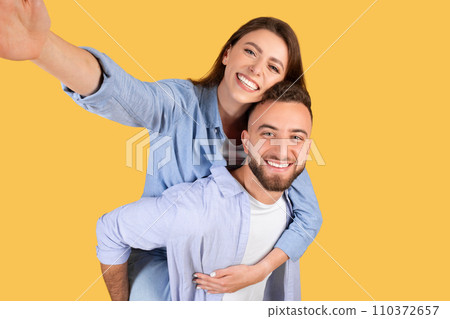 Happy loving european couple taking selfie together, cheerful young woman piggybacking her boyfriend, yellow background 110372657