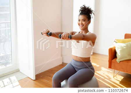 Fit woman sitting on balance ball lifting weights exercising indoor 110372674