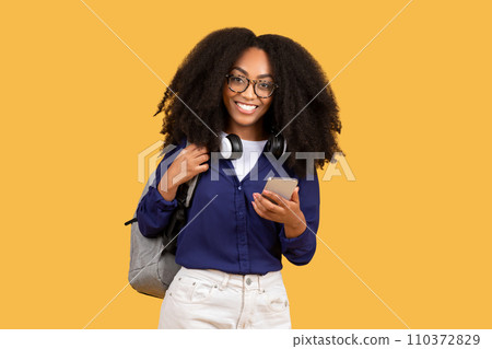 Excited black lady student in glasses with backpack, using cellphone and smiling, on yellow background Excited black lady student in glasses with backpack, using cellphone and smiling, on yellow background 110372829