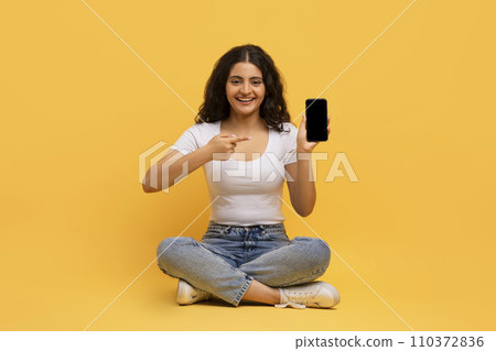Cheerful attractive young hindu lady with smartphone sitting on floor Cheerful attractive young hindu lady with smartphone sitting on floor 110372836