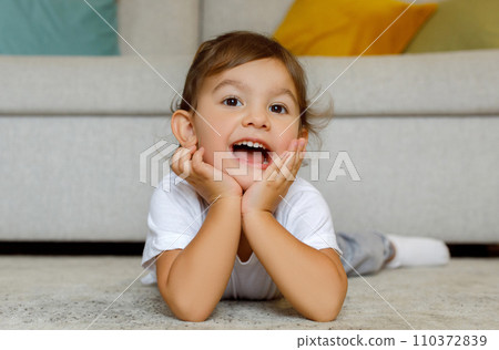 Adorable toddler girl in white t-shirt lying on floor, looking at camera 110372839