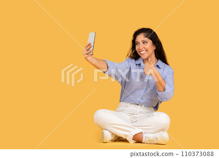 A cheerful young woman in casual clothes sits cross-legged on a yellow background A cheerful young woman in casual clothes sits cross-legged on a yellow background 110373088