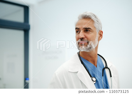 Portrait of confident mature doctor standing in Hospital corridor. Handsome doctor with gray hair wearing white coat, stethoscope around neck standing in modern private clinic, looking at camera. Portrait of confident mature doctor standing in Hospital corridor. Handsome doctor with gray hair wearing white coat, stethoscope around neck standing in modern private clinic, looking at camera. 110373125