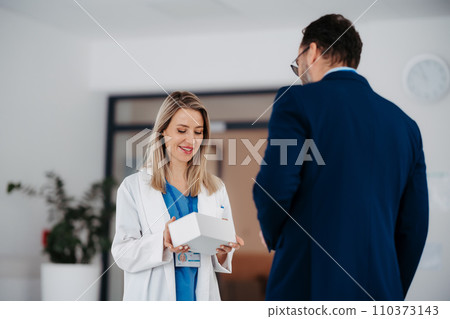 Pharmaceutical sales representative talking with female doctor in medical building, convicing her to try new pharmaceutical product. Doctor holding box with drug samples. 110373143