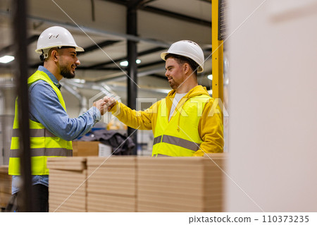 Young man with Down syndrome working in modern factory, fist bump with colleague, teamwork. Concept of workers with disabilities, support in workplace. 110373235
