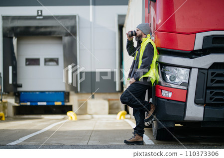 Truck driver leaning against red truck and drinking coffee, waiting for warehouse workers. 110373306