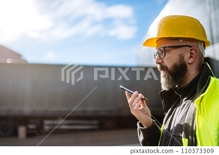 Warehouse worker standing outdoors, phone calling with truck driver. Warehouse receiver waiting for a delivery. 110373309
