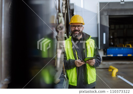 Portrait of smiling warehouse receiver standing by blue truck and holding tablet. Receiving clerk looking at cargo details, checking delivered items or goods against order. 110373322