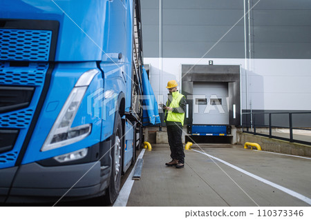 Warehouse receiver standing by blue truck, holding tablet, looking at cargo details, checking delivered items, goods against order. Warehouse receiver standing by blue truck, holding tablet, looking at cargo details, checking delivered items, goods against order. 110373346