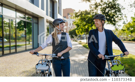 Spouses commuting through the city, talking and walking by bike on street. Middle-aged city commuters traveling from work by bike after a long workday. 110373348