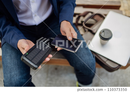 Businessman, freelancer or manager working outdoors in city park. Man with laptop on knees drinking coffee, charging his smartphone with solar phone charger. Concept of working remotely. 110373350