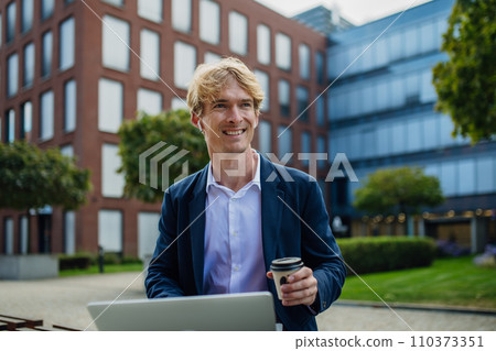 Businessman, freelancer or manager working outdoors in city park. Man with laptop on knees drinking coffee, having video call, listening music. Concept of working remotely. 110373351