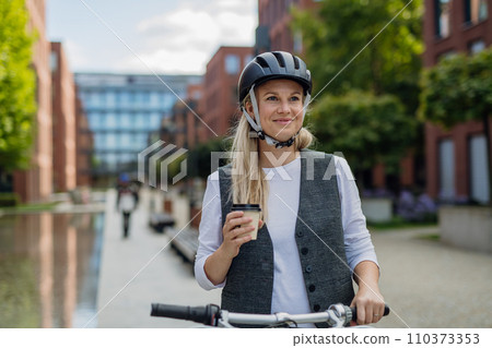 Beautiful middle-aged woman commuting through the city, buying, drinking coffe in front of office. Female city commuter traveling from work by bike after a long workday. 110373353