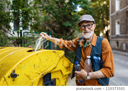 Senior man throwing plastic waste, bottles into recycling container in front apartment. Elderly man sorting the waste according to material into colored bins. 110373501