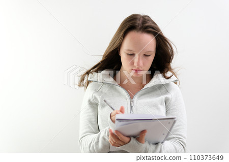 Portrait of attractive optimistic woman wearing sports suit standing with paper notebook and pen in hands, looking at camera with smile. Indoor studio shot isolated on gray background. 110373649