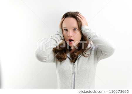 Portrait of shocked young Caucasian woman wearing white shirt holding head in hands and looking at camera. Fear and surprise concept 110373655