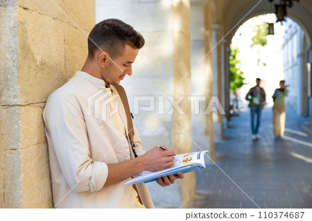 Handsome young man standing near the wall with a textbook in hands Handsome young man standing near the wall with a textbook in hands 110374687
