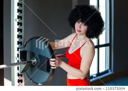 A woman with curly hair puts an iron wheel on a sports barbell A woman with curly hair puts an iron wheel on a sports barbell 110375004