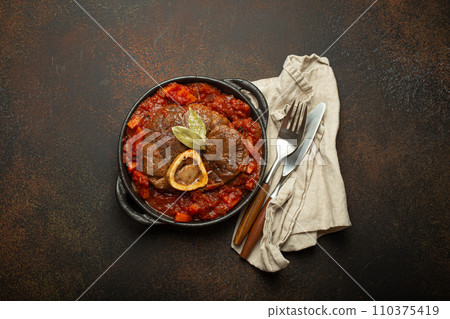 Traditional Italian dish Ossobuco all Milanese made with cut veal shank meat with vegetable tomato sauce served in black casserole pan top view on rustic brown background 110375419