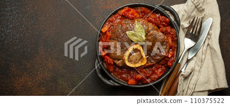 Traditional Italian dish Ossobuco all Milanese made with cut veal shank meat with vegetable tomato sauce served in black casserole pan top view on rustic brown background 110375522