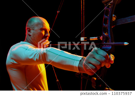 Portrait of serious man, archer aiming with bow on target against black studio background in neon light. Archery sport, competition Portrait of serious man, archer aiming with bow on target against black studio background in neon light. Archery sport, competition 110375937