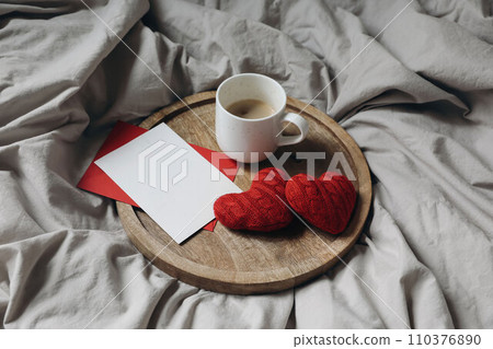Love, engagement concept. Two red knitted hearts, cup of coffee on wooden plate. Breakfast in bed. Linen duvet cover, bedding. Empty Valentines day greeting card, invitation. Flatlay, top view, 110376890