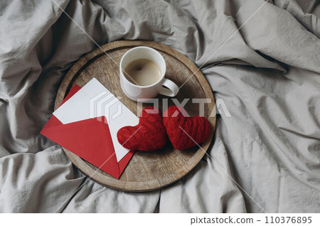Valentines day still life. Blank greeting card, invitation mockup, red envelope on wooden tray. Couple of red textile hearts. Cup of coffee in bed. Linen duvet cover. Love concept. Flatlay, top view. 110376895
