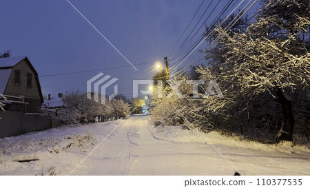 Snowy country road at evening time. Street lamp illuminating snow trail at winter night. Beautiful nature landscape with falling snowflakes at background. Snowy country road at evening time. Street lamp illuminating snow trail at winter night. Beautiful nature landscape with falling snowflakes at background. 110377535