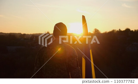 Male ukrainian army soldier stands with national banner against background of beautiful sunset. Young man in camouflage uniform with Ukraine flag as symbol of victory against russian aggression Male ukrainian army soldier stands with national banner against background of beautiful sunset. Young man in camouflage uniform with Ukraine flag as symbol of victory against russian aggression 110377782