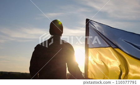 Ukrainian army man stands with national banner at countryside on sunset. Male soldier in military uniform with Ukraine flag as symbol of victory against russian aggression. Invasion resistance concept Ukrainian army man stands with national banner at countryside on sunset. Male soldier in military uniform with Ukraine flag as symbol of victory against russian aggression. Invasion resistance concept 110377791