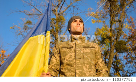 Ukrainian army man stands with national banner at countryside against blue sky. Male soldier in military uniform with Ukraine flag as symbol of victory against russian aggression. Invasion resistance. 110377792