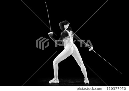 Striking profile of two-sword-wielding sportswoman, radiating power and grace, athleticism and dedication against black studio background. 110377950