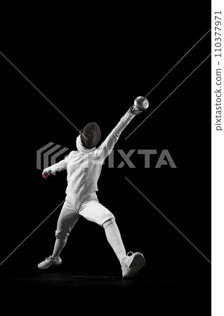 Dynamic portrait of female fencer lunges forward, her blade poised to strike, she closing in fast against black studio background. Dynamic portrait of female fencer lunges forward, her blade poised to strike, she closing in fast against black studio background. 110377971