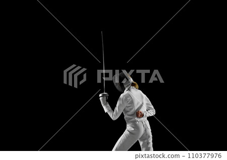Female fencer stands at ready, her blade held high, poised to strike, but she waiting, her opponent's every move calculated against black studio background. Female fencer stands at ready, her blade held high, poised to strike, but she waiting, her opponent's every move calculated against black studio background. 110377976