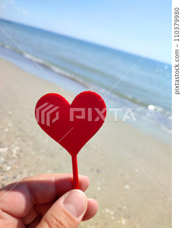 Person holding in fingers hand stick in shape red heart on background sea and sea waves, blue sky in seashore on sunny summer day close-up. Concept love romance amour St Valentines Day Person holding in fingers hand stick in shape red heart on background sea and sea waves, blue sky in seashore on sunny summer day close-up. Concept love romance amour St Valentines Day 110379980