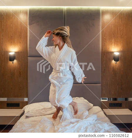 Woman having fun and jumping with bathrobe on bed in hotel room Woman having fun and jumping with bathrobe on bed in hotel room 110382612