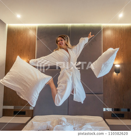 Woman having fun and jumping with bathrobe on bed in hotel room Woman having fun and jumping with bathrobe on bed in hotel room 110382620